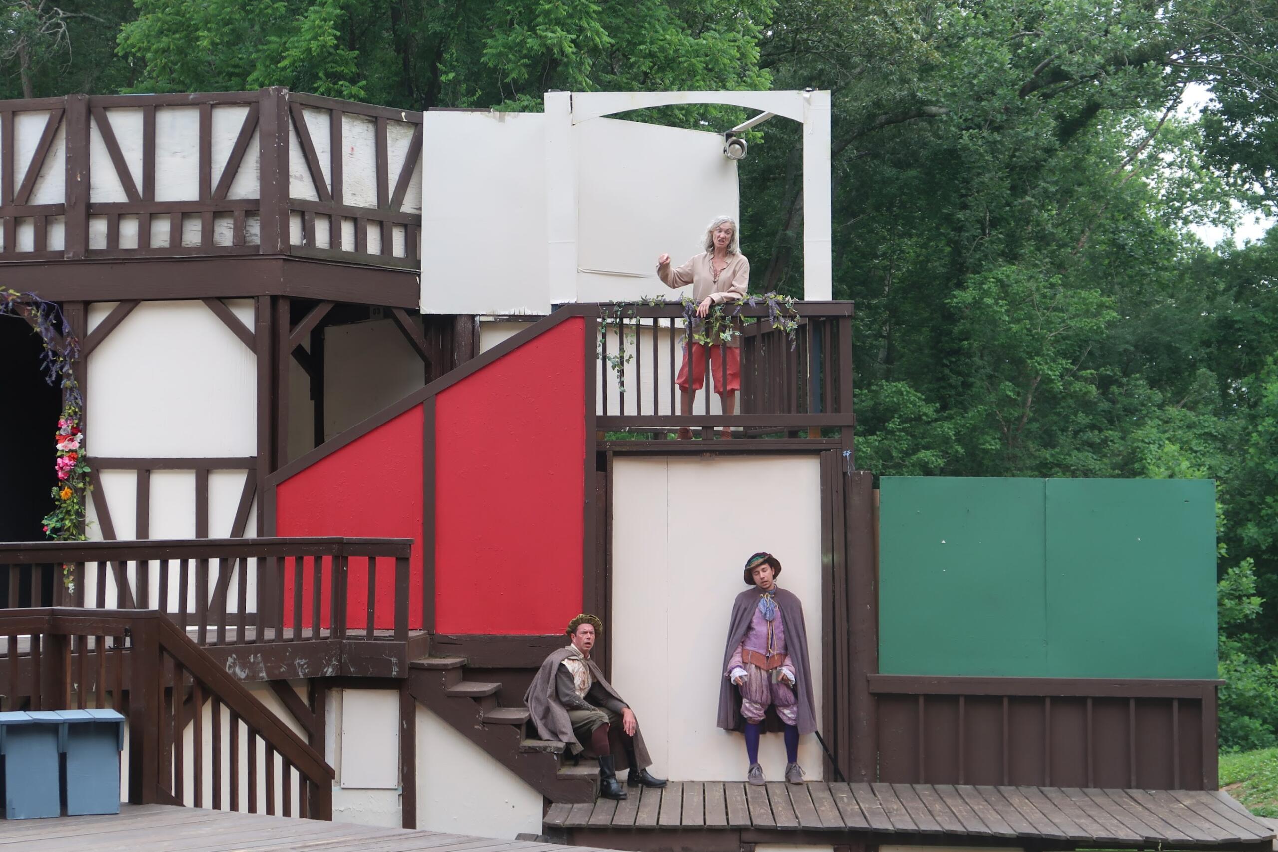 Actors in period costumes stand on a multi-level wooden stage with red and white panels, greenery in the background, resembling an outdoor theater.