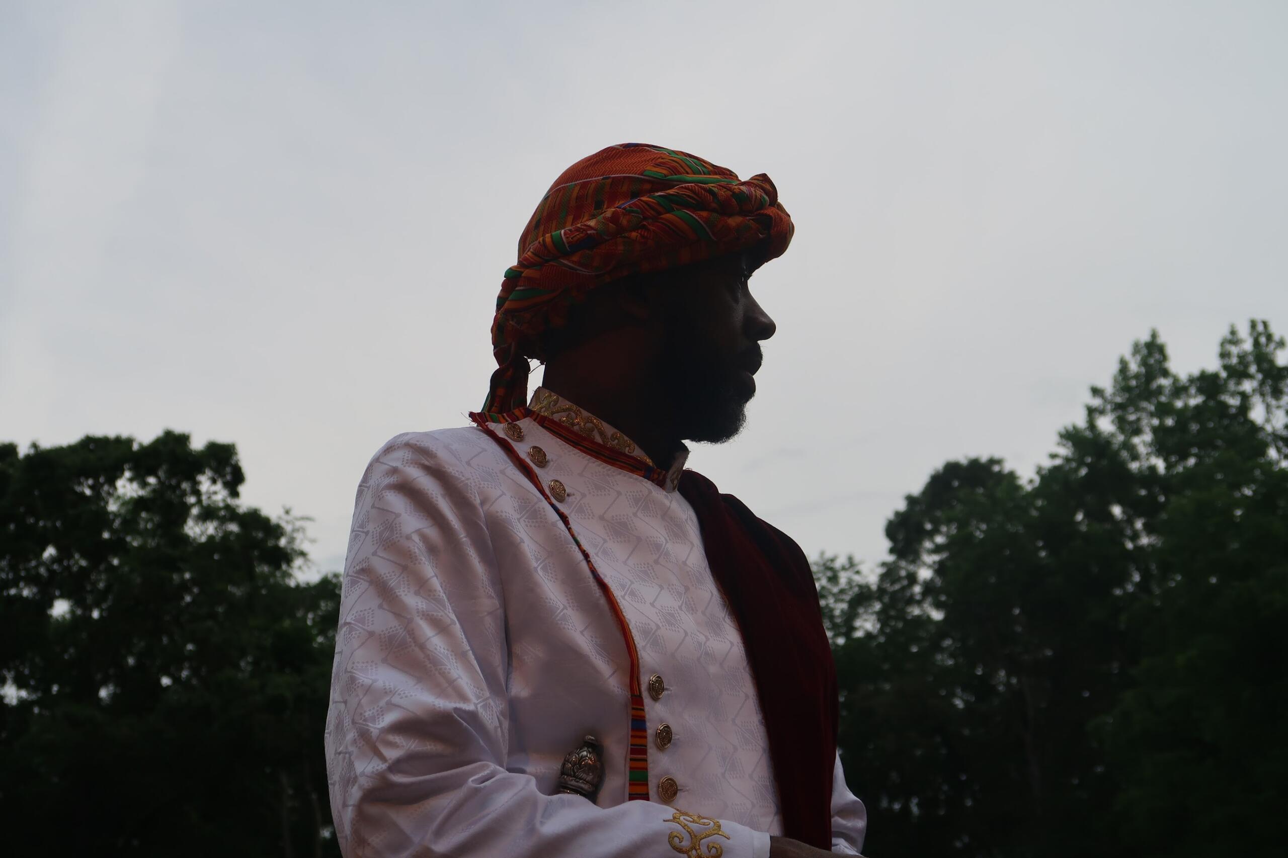 Profile silhouette of a man in a colorful patterned turban and white embroidered shirt outdoors with trees in the background
