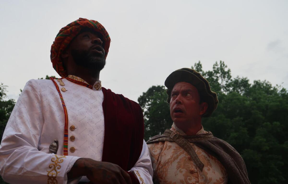 Two men in ornate historical costumes stand outdoors, looking upward toward the sky; one wears a white embroidered tunic with gold buttons and a red and green turban, the other a beige brocade outfit with a brown cloak and hat.