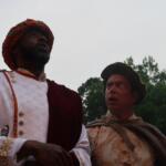 Two men in ornate historical costumes stand outdoors, looking upward toward the sky; one wears a white embroidered tunic with gold buttons and a red and green turban, the other a beige brocade outfit with a brown cloak and hat.