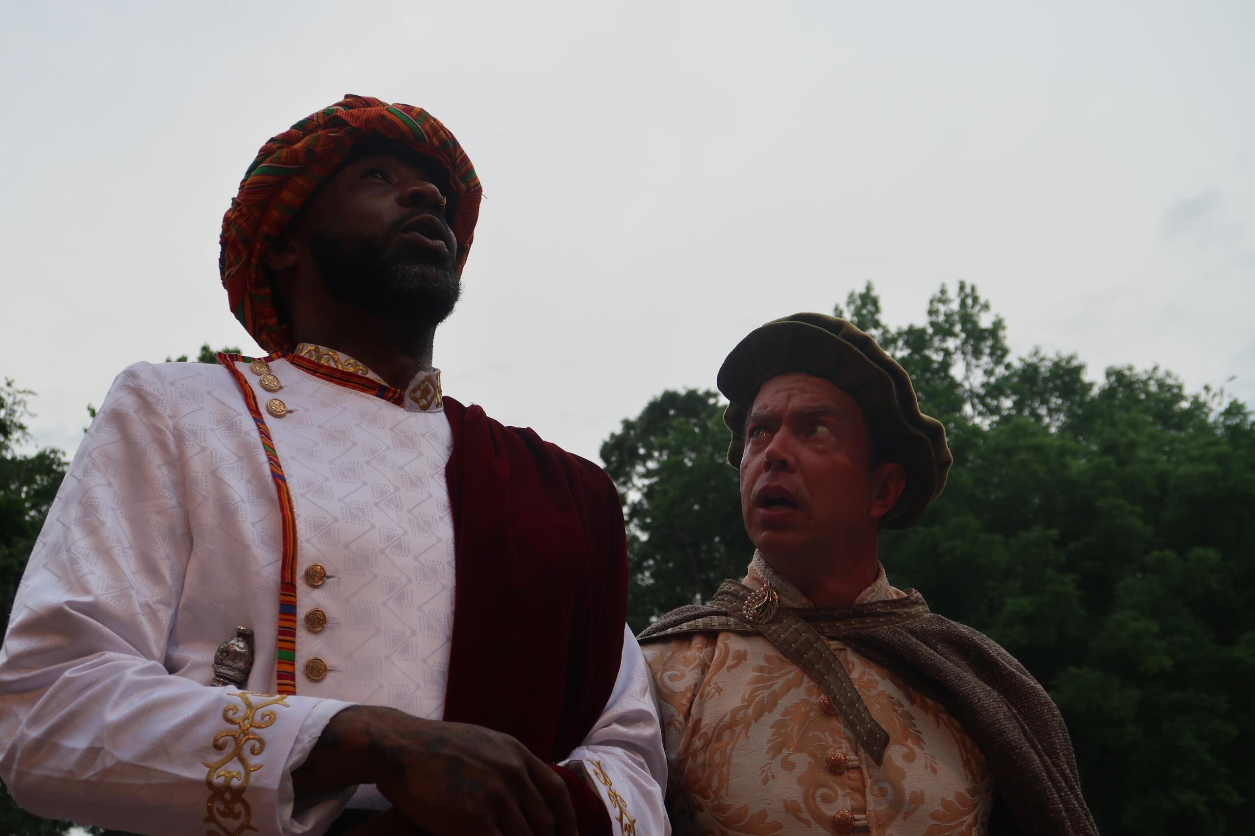 Two men in ornate historical costumes stand outdoors, looking upward toward the sky; one wears a white embroidered tunic with gold buttons and a red and green turban, the other a beige brocade outfit with a brown cloak and hat.