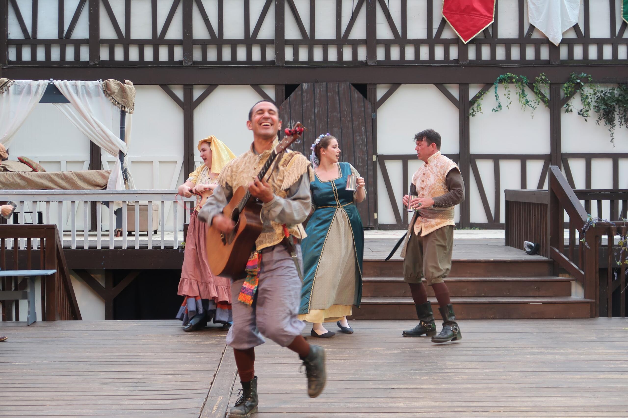Group of performers in period costumes on a wooden stage; a man plays guitar and others hold cups, mid-performance on a Tudor-style set.