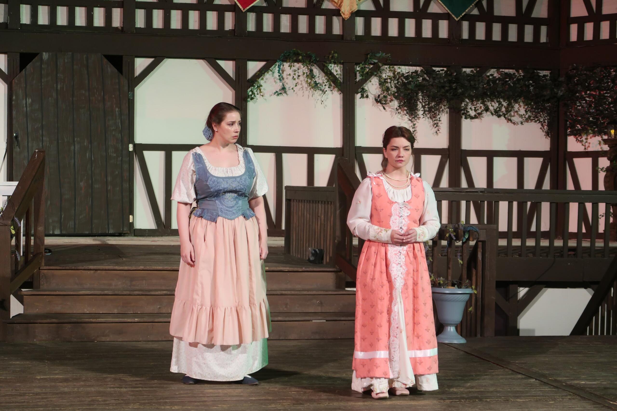 Two women in period dresses stand on a wooden stage with a Tudor-style backdrop.