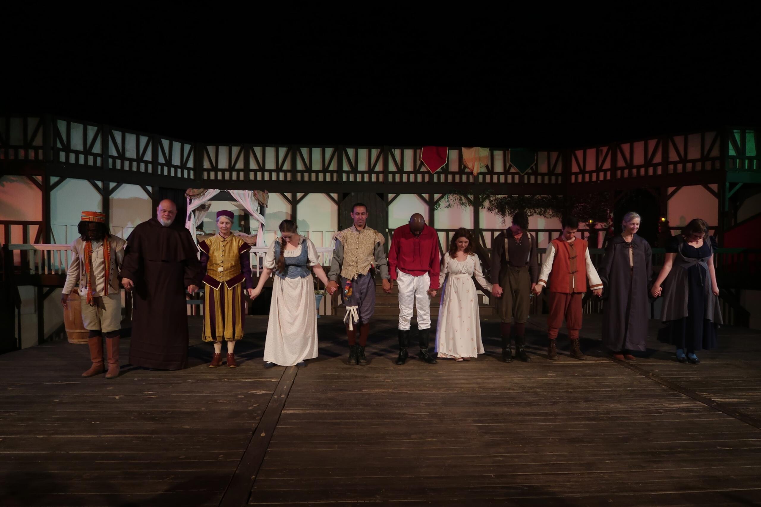 Cast of performers in medieval costumes holding hands on a stage during a curtain call or final bow.