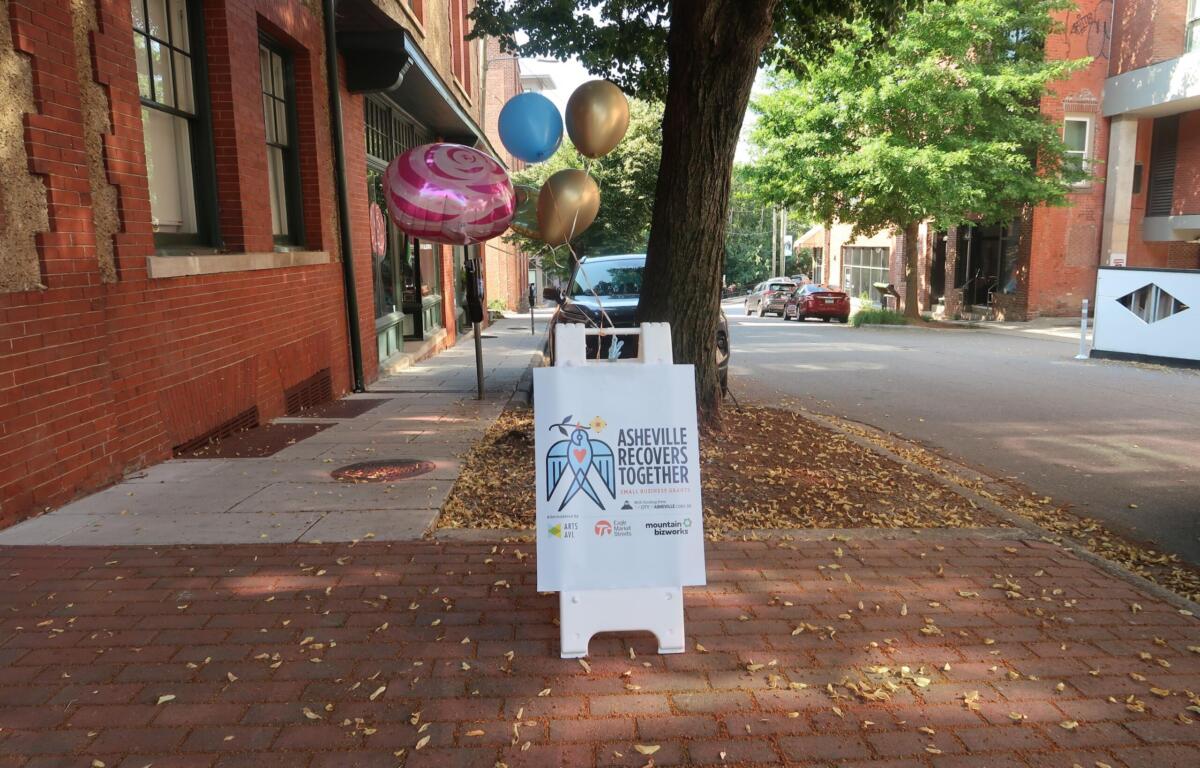 Balloon cluster tied to a post on a brick sidewalk beside a brick building, with a sign reading Asheville Recovers Together nearby.