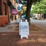Balloon cluster tied to a post on a brick sidewalk beside a brick building, with a sign reading Asheville Recovers Together nearby.