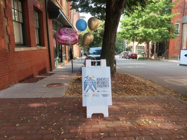 Balloon cluster tied to a post on a brick sidewalk beside a brick building, with a sign reading Asheville Recovers Together nearby.