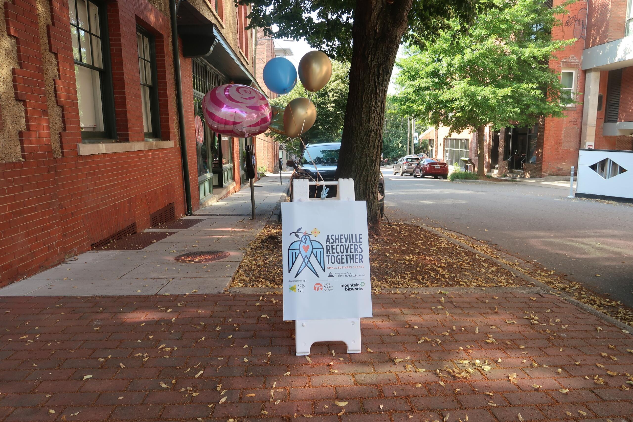 Balloon cluster tied to a post on a brick sidewalk beside a brick building, with a sign reading Asheville Recovers Together nearby.