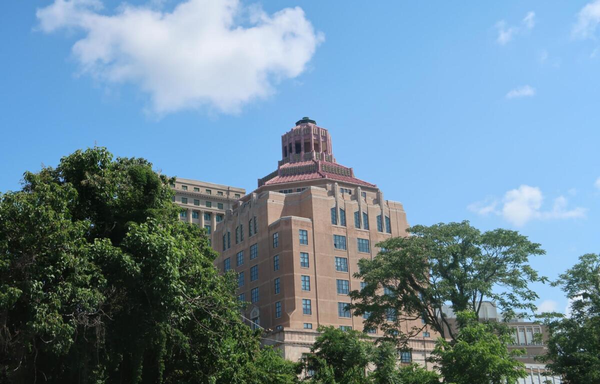 Tall brick building with a decorative tiered rooftop rising above green trees against a blue sky.