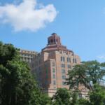 Tall brick building with a decorative tiered rooftop rising above green trees against a blue sky.
