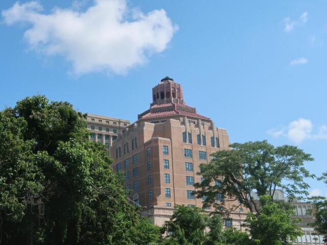 Tall brick building with a decorative tiered rooftop rising above green trees against a blue sky.