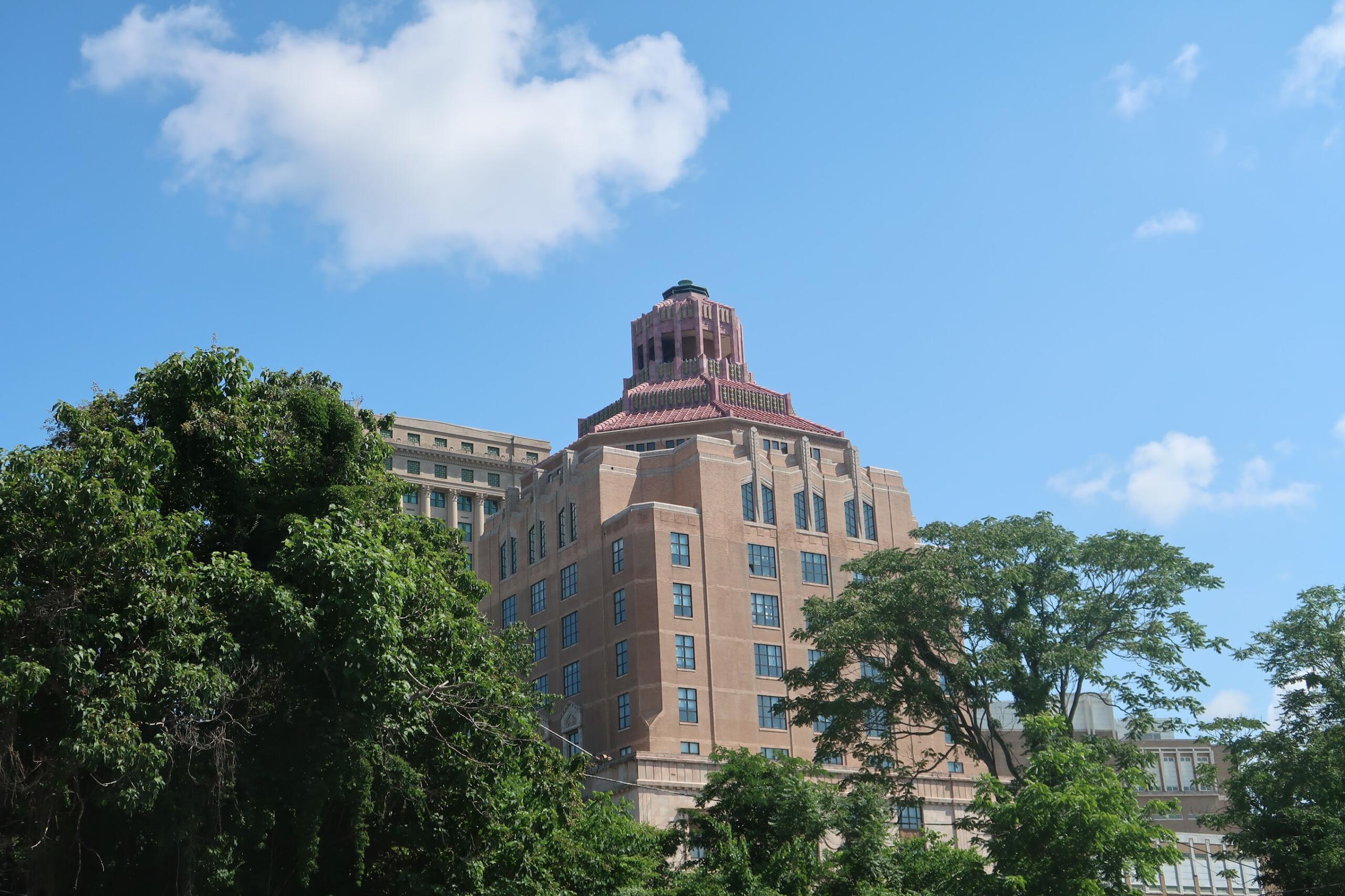 Tall brick building with a decorative tiered rooftop rising above green trees against a blue sky.
