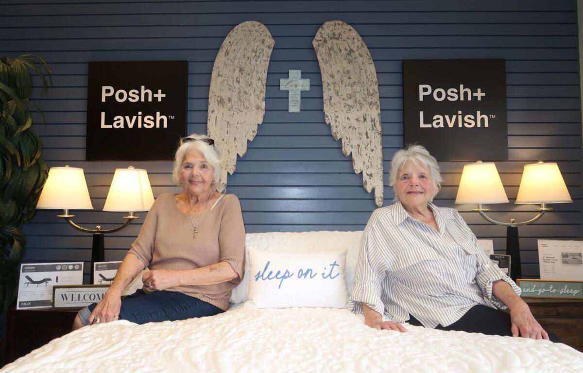 Two elderly women sit on a white quilt in a showroom, with blue paneled wall, angel wing art and a cross between them, flanked by Posh+ Lavish signs and lamps on both sides