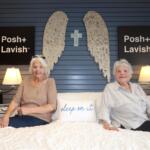 Two elderly women sit on a white quilt in a showroom, with blue paneled wall, angel wing art and a cross between them, flanked by Posh+ Lavish signs and lamps on both sides