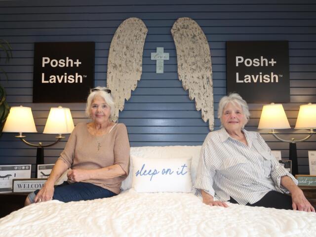 Two elderly women sit on a white quilt in a showroom, with blue paneled wall, angel wing art and a cross between them, flanked by Posh+ Lavish signs and lamps on both sides