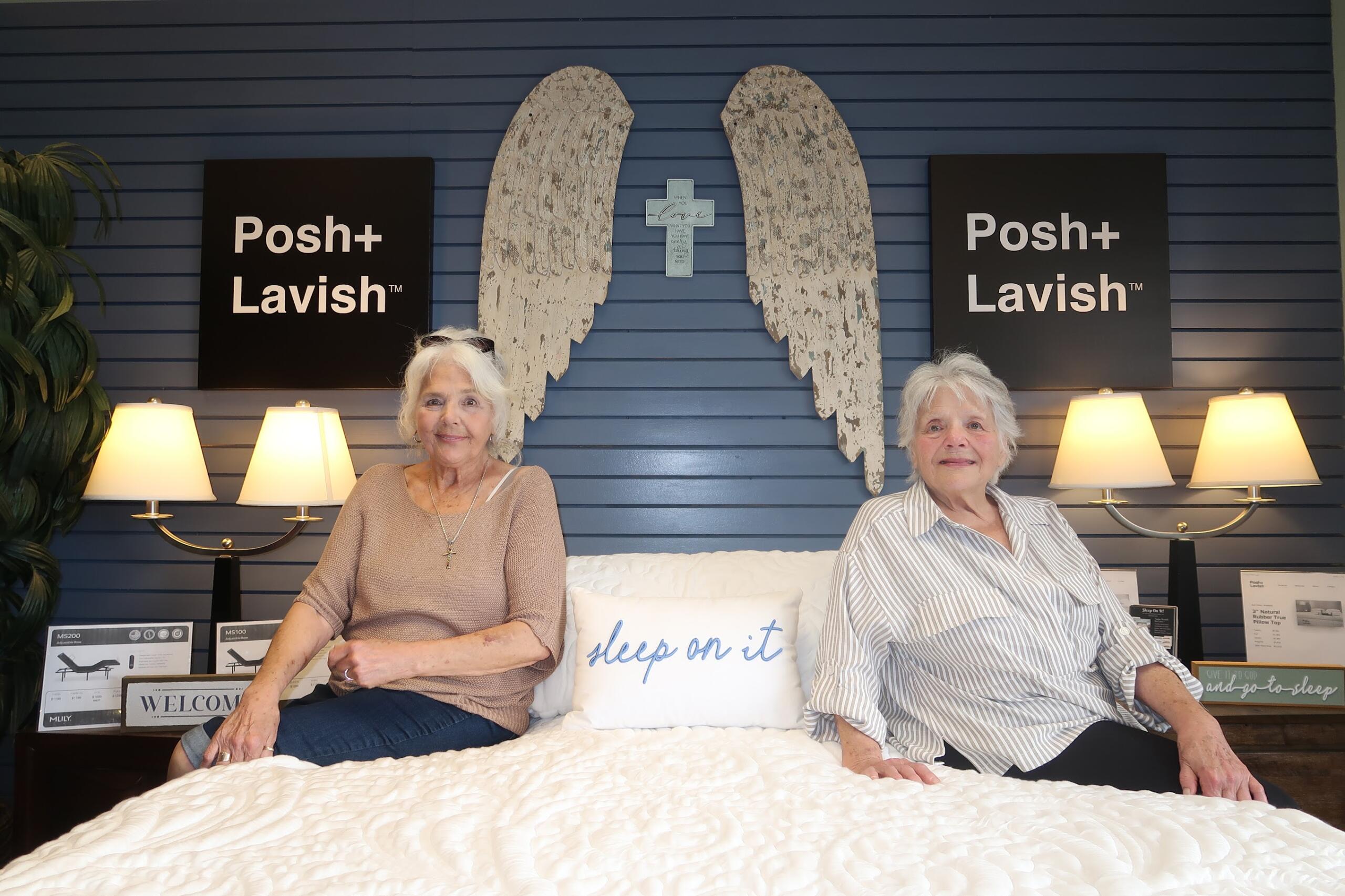 Two elderly women sit on a white quilt in a showroom, with blue paneled wall, angel wing art and a cross between them, flanked by Posh+ Lavish signs and lamps on both sides