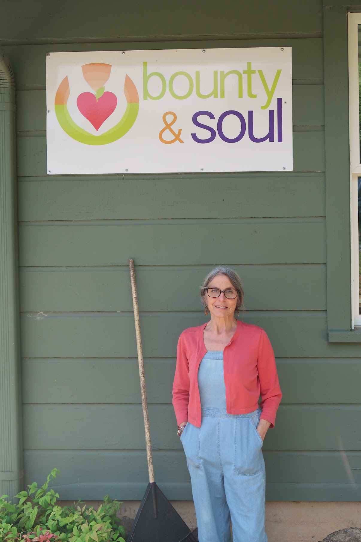 Woman in pink cardigan and light-blue overalls stands next to a 'bounty & soul' sign on a green building, holding a long-handled rake outside.