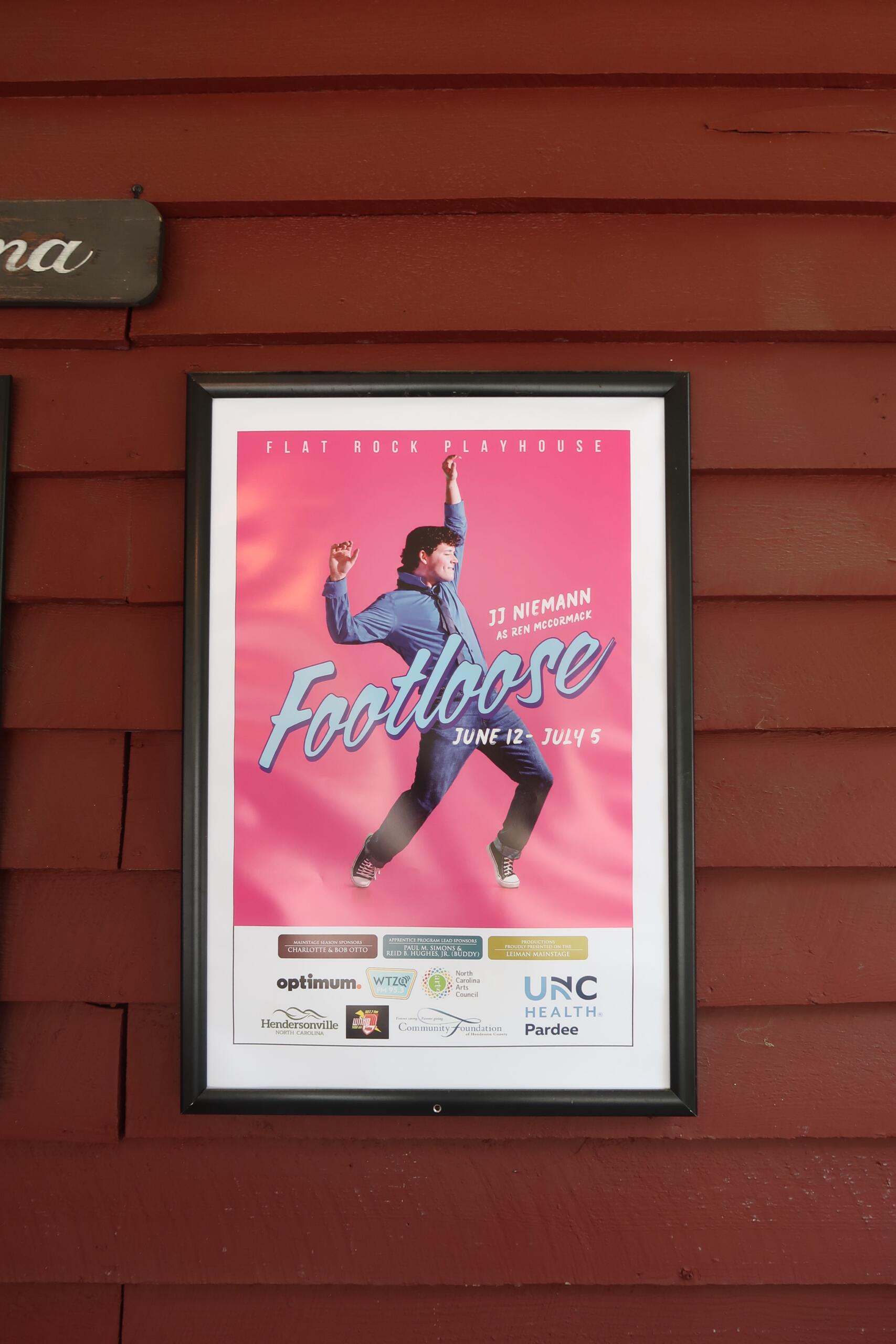 Framed Footloose theater poster with a dancer leaping on a pink background, mounted on a red wooden wall.