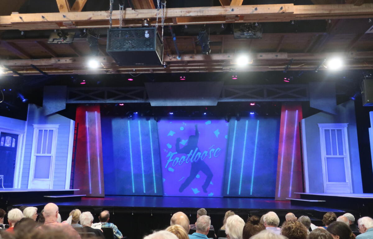 Theater stage with a blue-lit backdrop displaying 'Footloose' and silhouettes; audience seated in the foreground.