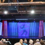 Theater stage with a blue-lit backdrop displaying 'Footloose' and silhouettes; audience seated in the foreground.