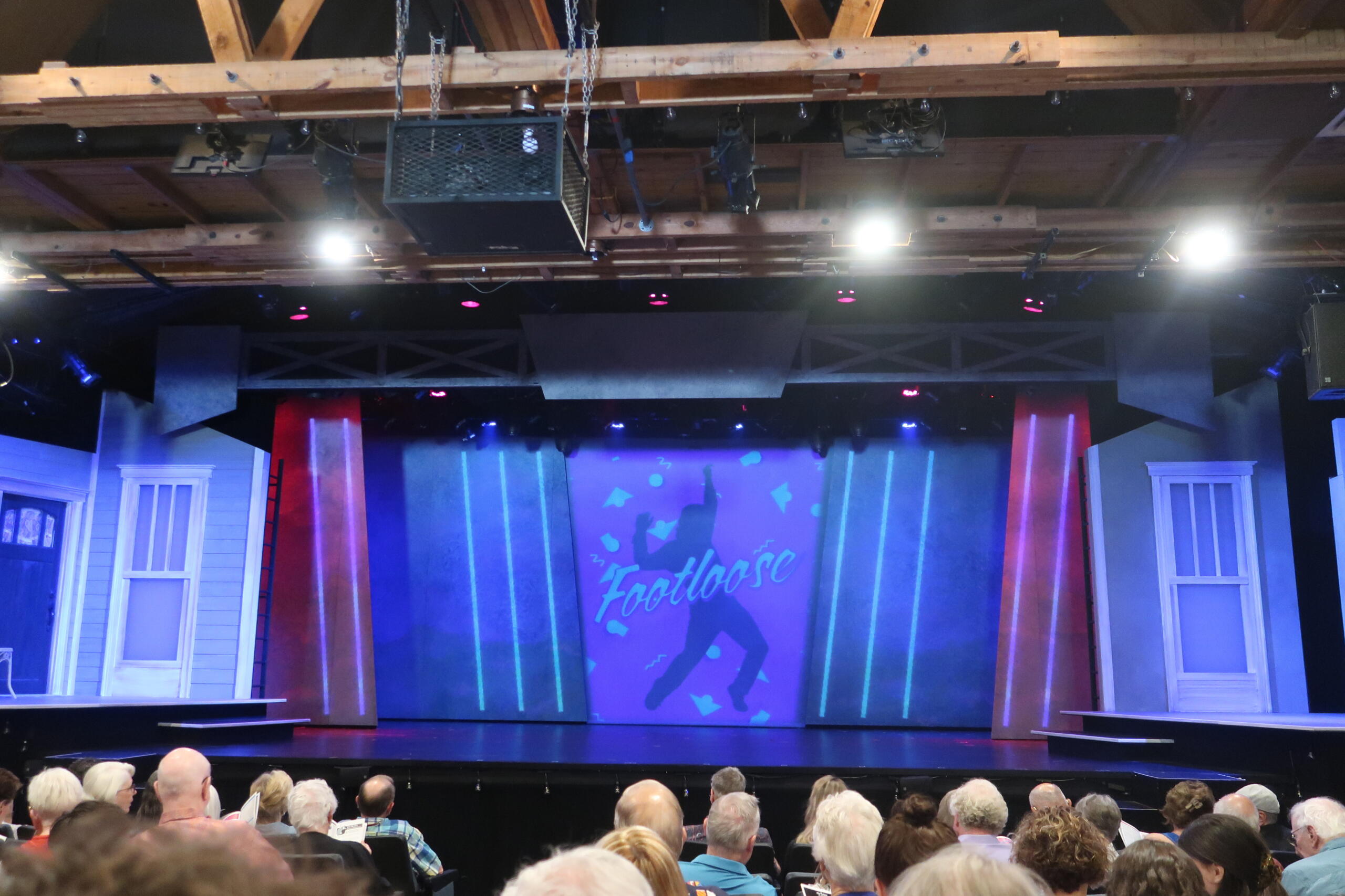 Theater stage with a blue-lit backdrop displaying 'Footloose' and silhouettes; audience seated in the foreground.