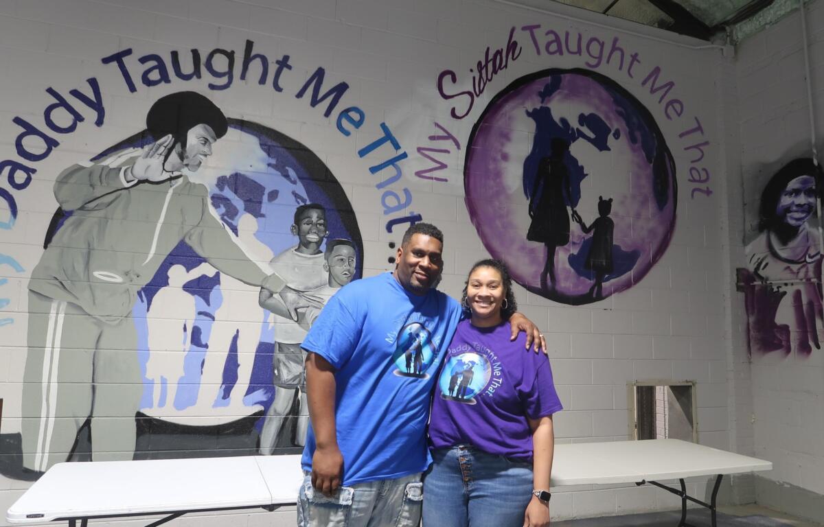 Two people stand smiling in front of a mural with a purple globe and inspirational text on the wall behind them.