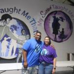 Two people stand smiling in front of a mural with a purple globe and inspirational text on the wall behind them.