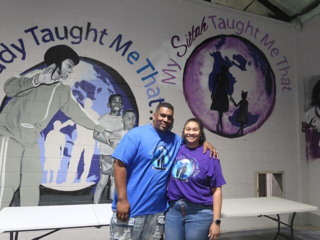 Two people stand smiling in front of a mural with a purple globe and inspirational text on the wall behind them.