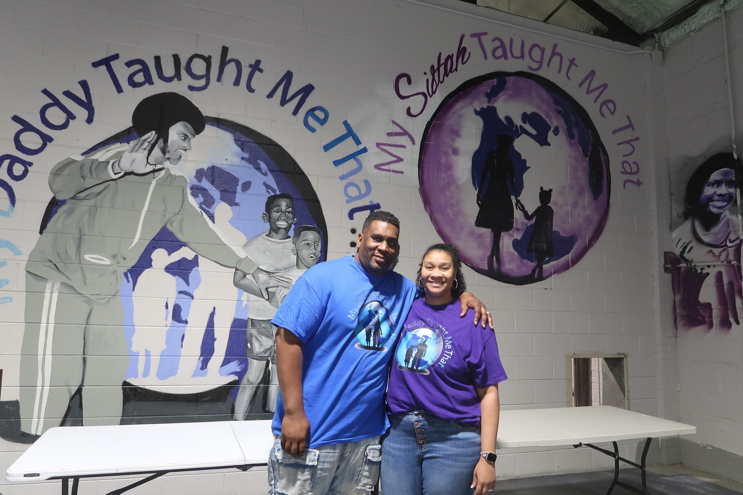 Two people stand smiling in front of a mural with a purple globe and inspirational text on the wall behind them.