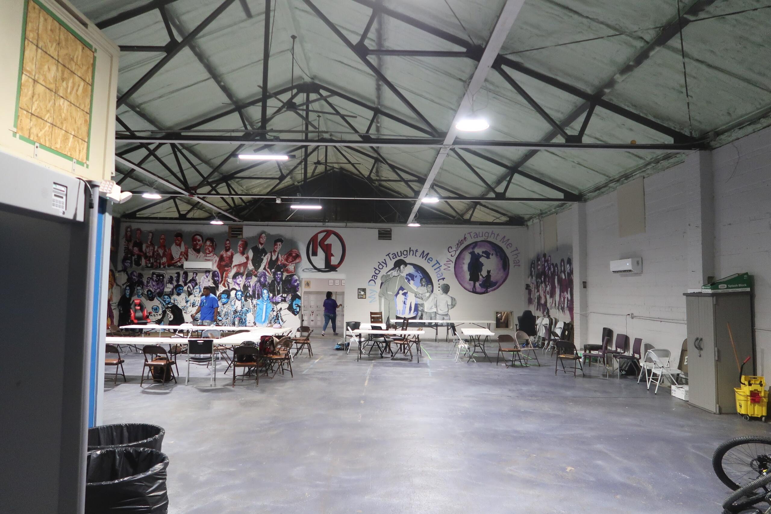 Industrial interior with exposed steel trusses, bright overhead lights, and scattered folding tables and chairs across a large open space.