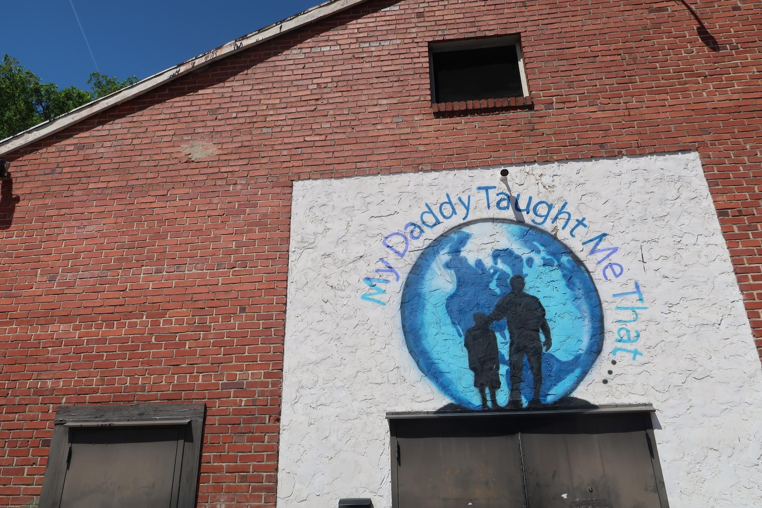 Brick red wall with a large white mural showing blue circle and two silhouetted figures, surrounded by curved blue text. A small dark window is above the mural.