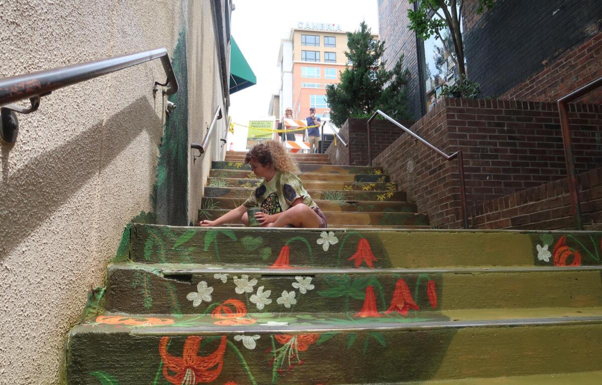 Person crouching on graffiti-painted steps, holding a cup, in an urban alley with mural flowers and leaves. Two people stand at the top behind caution tape.