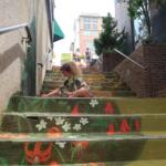 Person crouching on graffiti-painted steps, holding a cup, in an urban alley with mural flowers and leaves. Two people stand at the top behind caution tape.