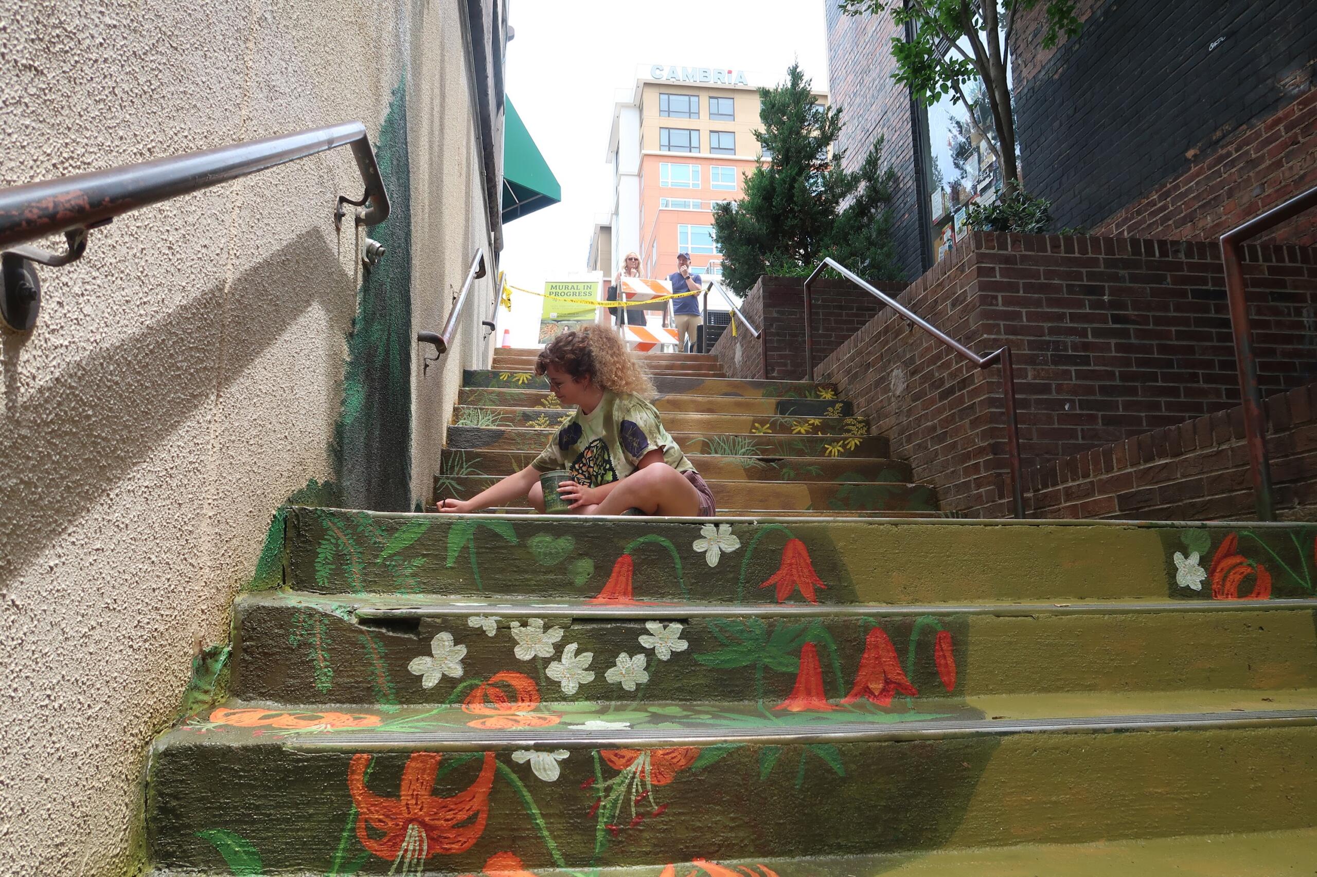 Person crouching on graffiti-painted steps, holding a cup, in an urban alley with mural flowers and leaves. Two people stand at the top behind caution tape.