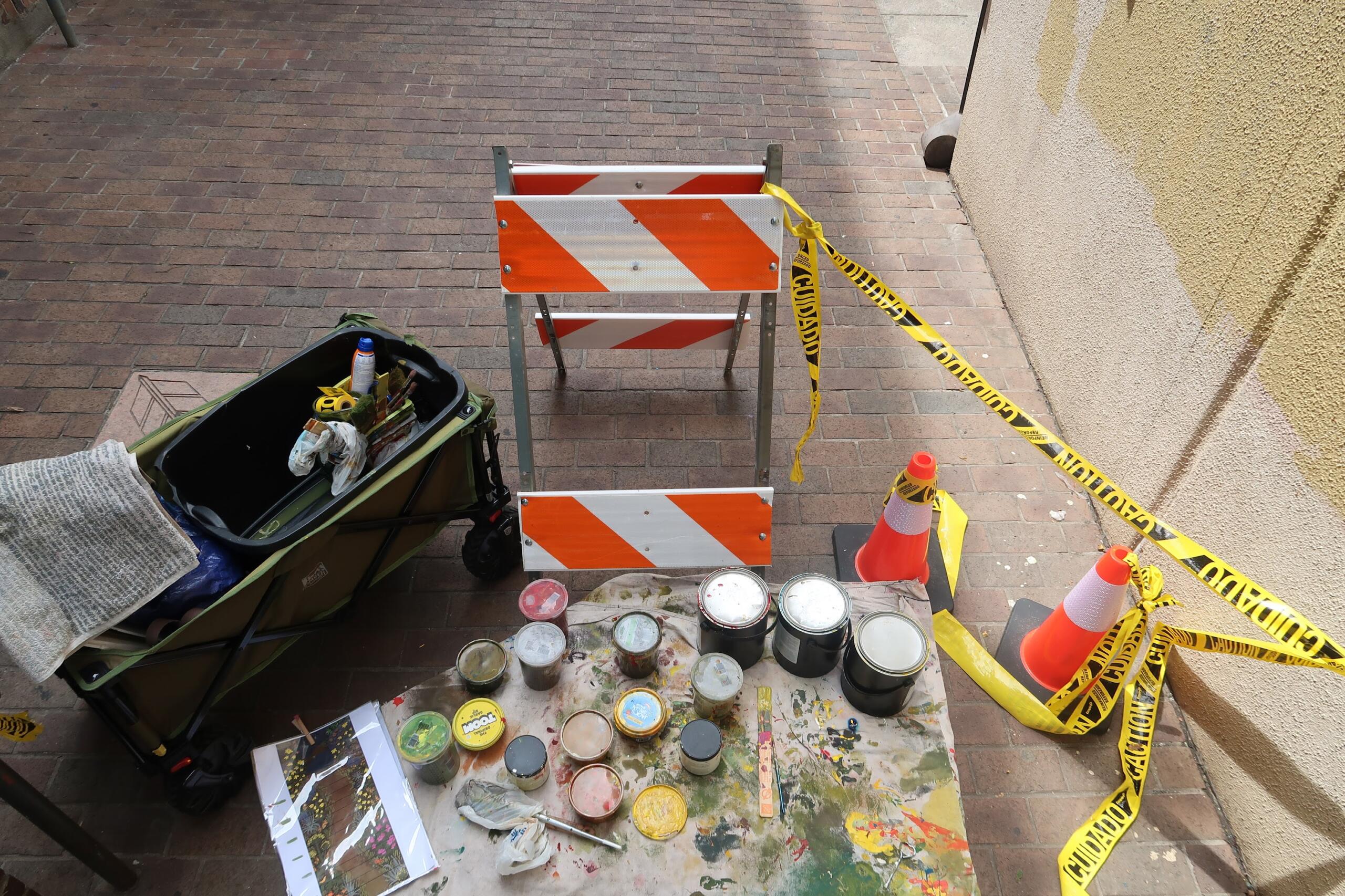 Construction barrier on a brick sidewalk with yellow caution tape and paint supplies nearby.