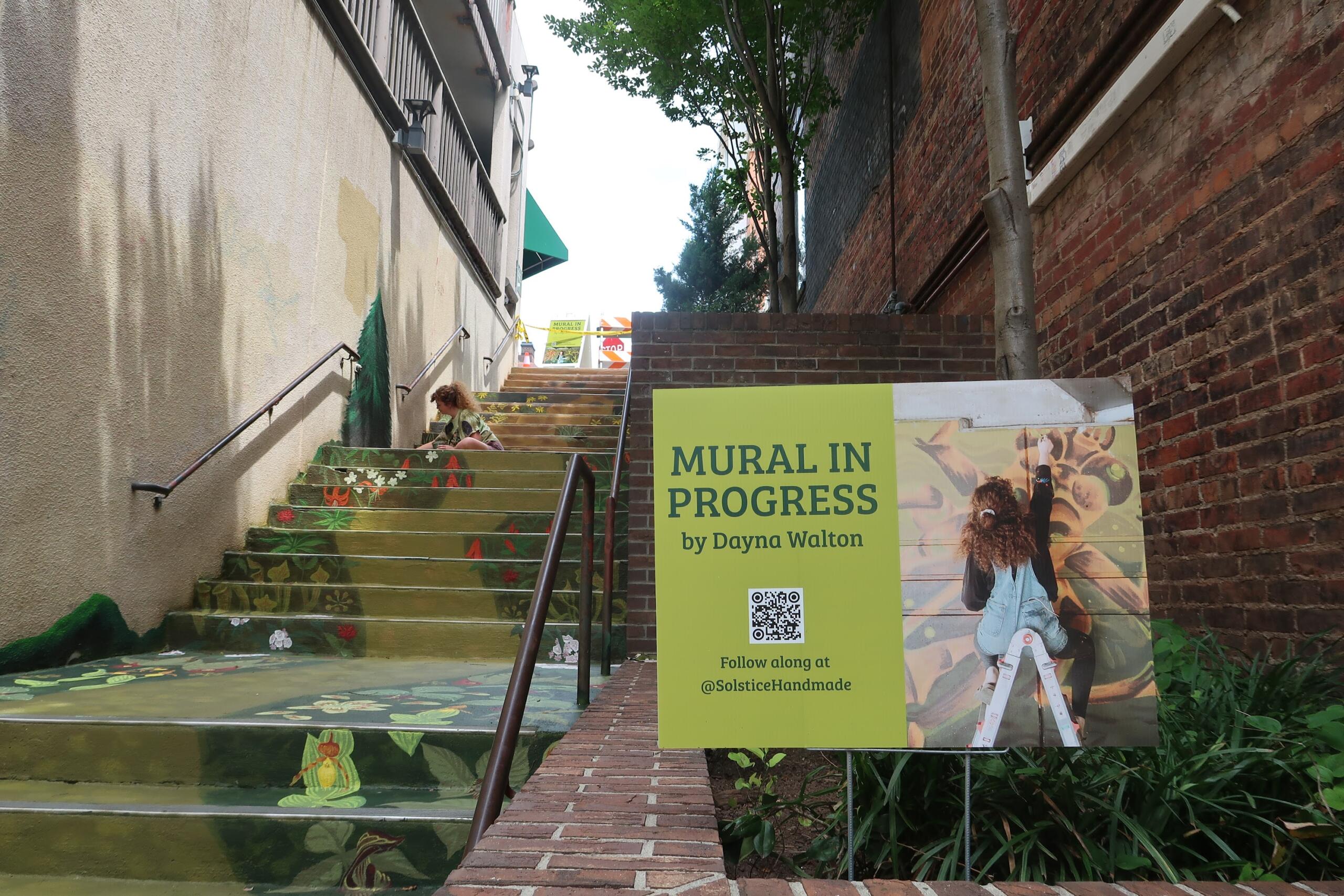 Brick stairway with a colorful mural on the risers and a large yellow sign reading 'MURAL IN PROGRESS' by Dayna Walton at the bottom.