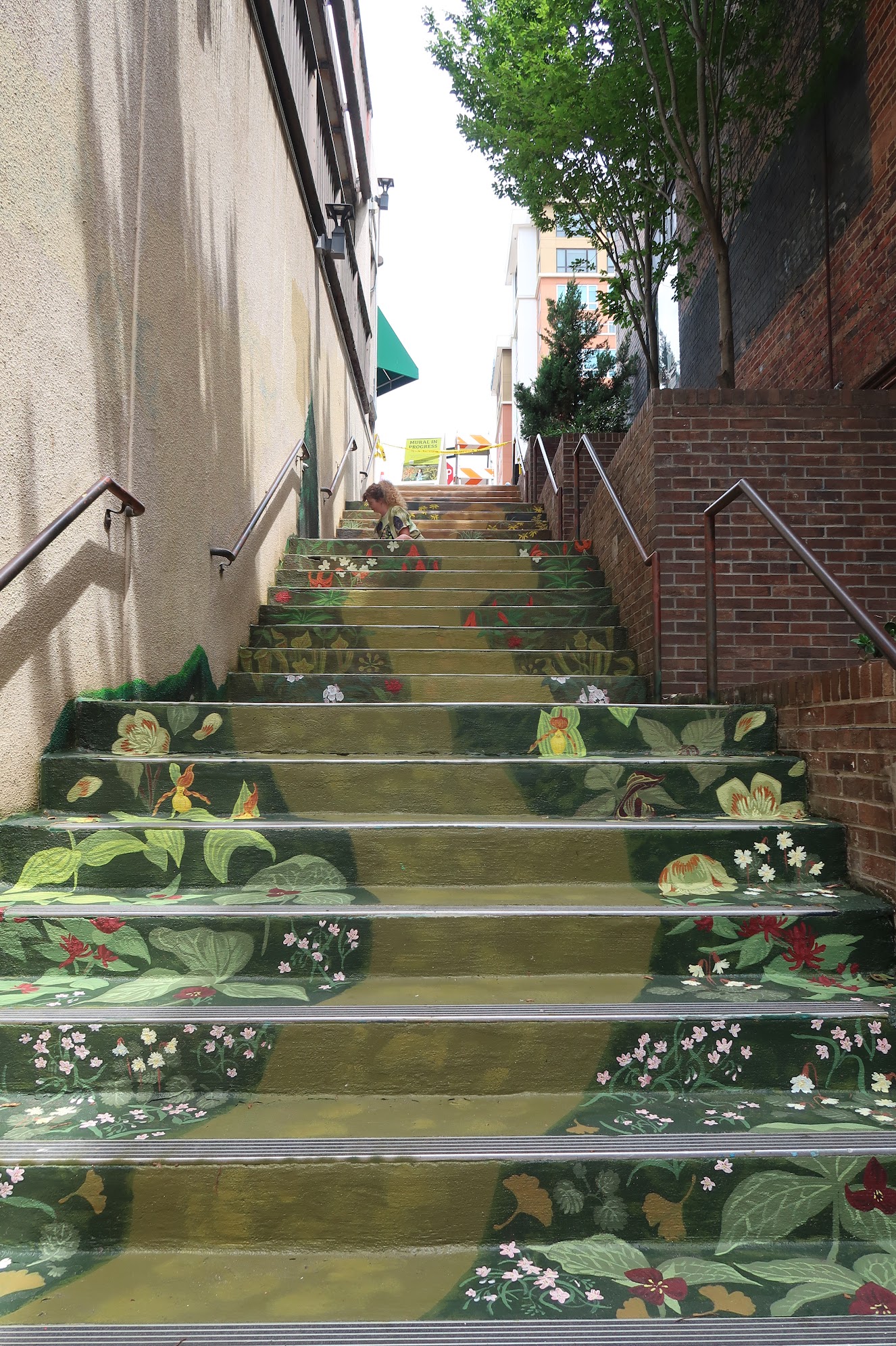 Outdoor stairway in an urban alley with green floral mural on each step; a person sits mid-steps amid plants and brick walls on either side.