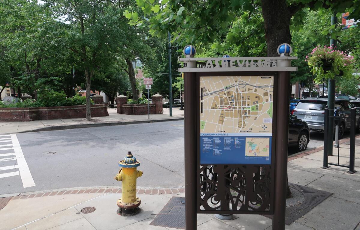 Informational map kiosk on a city sidewalk, surrounded by trees and parked cars in downtown Asheville.