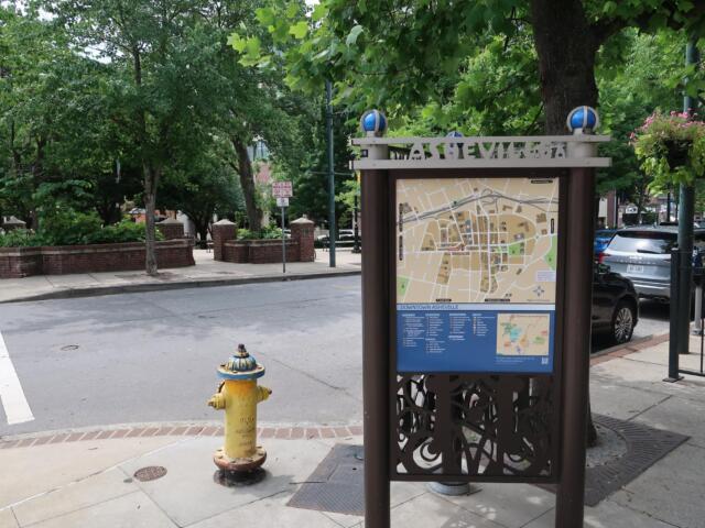 Informational map kiosk on a city sidewalk, surrounded by trees and parked cars in downtown Asheville.