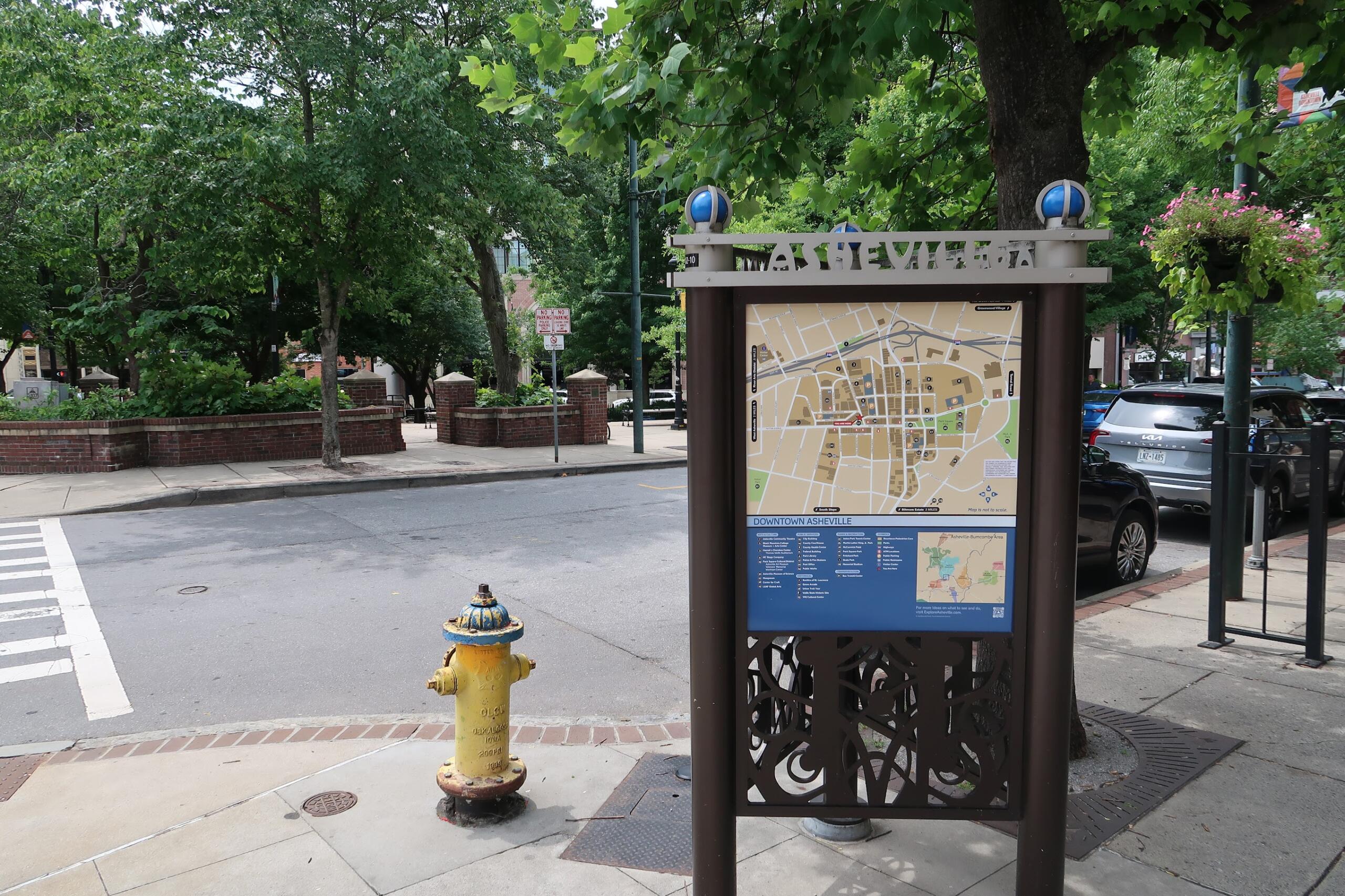 Informational map kiosk on a city sidewalk, surrounded by trees and parked cars in downtown Asheville.
