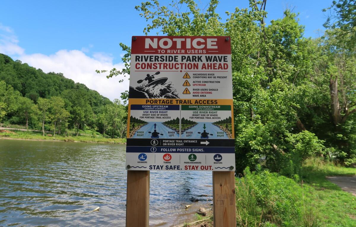 Sign near a river warning of Riverside Park wave construction ahead with logos and safety icons, trees in the background