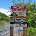 Sign near a river warning of Riverside Park wave construction ahead with logos and safety icons, trees in the background
