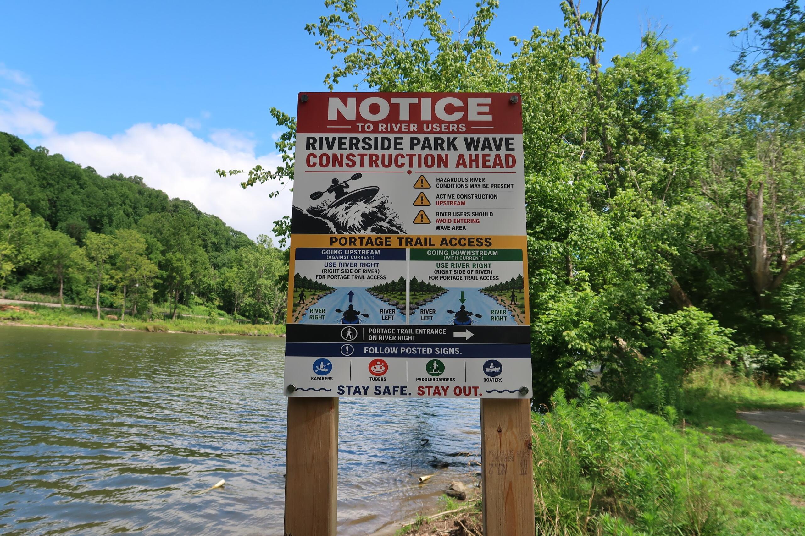 Sign near a river warning of Riverside Park wave construction ahead with logos and safety icons, trees in the background
