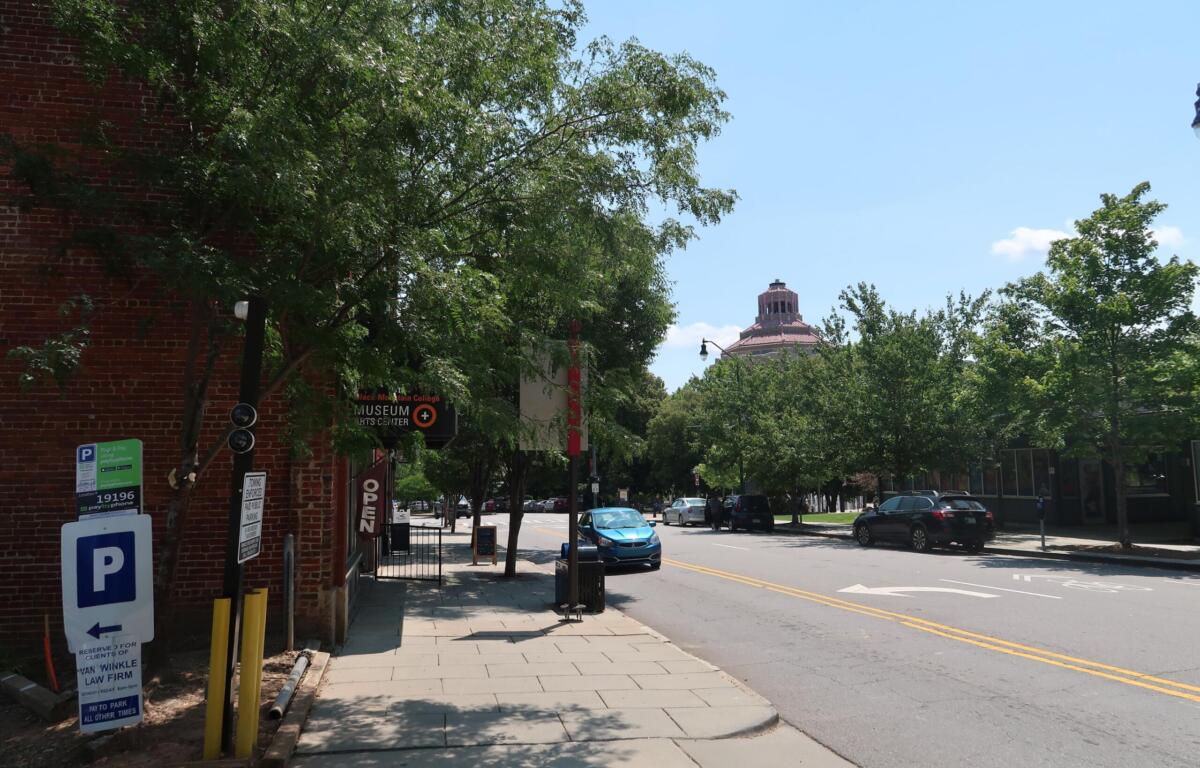 Street scene with a brick building and leafy trees, a sidewalk, and a distant domed building under a blue sky; cars parked along the street.