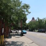 Street scene with a brick building and leafy trees, a sidewalk, and a distant domed building under a blue sky; cars parked along the street.