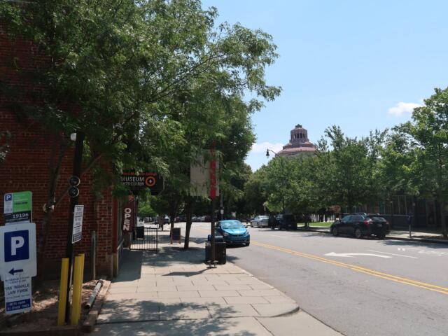 Street scene with a brick building and leafy trees, a sidewalk, and a distant domed building under a blue sky; cars parked along the street.