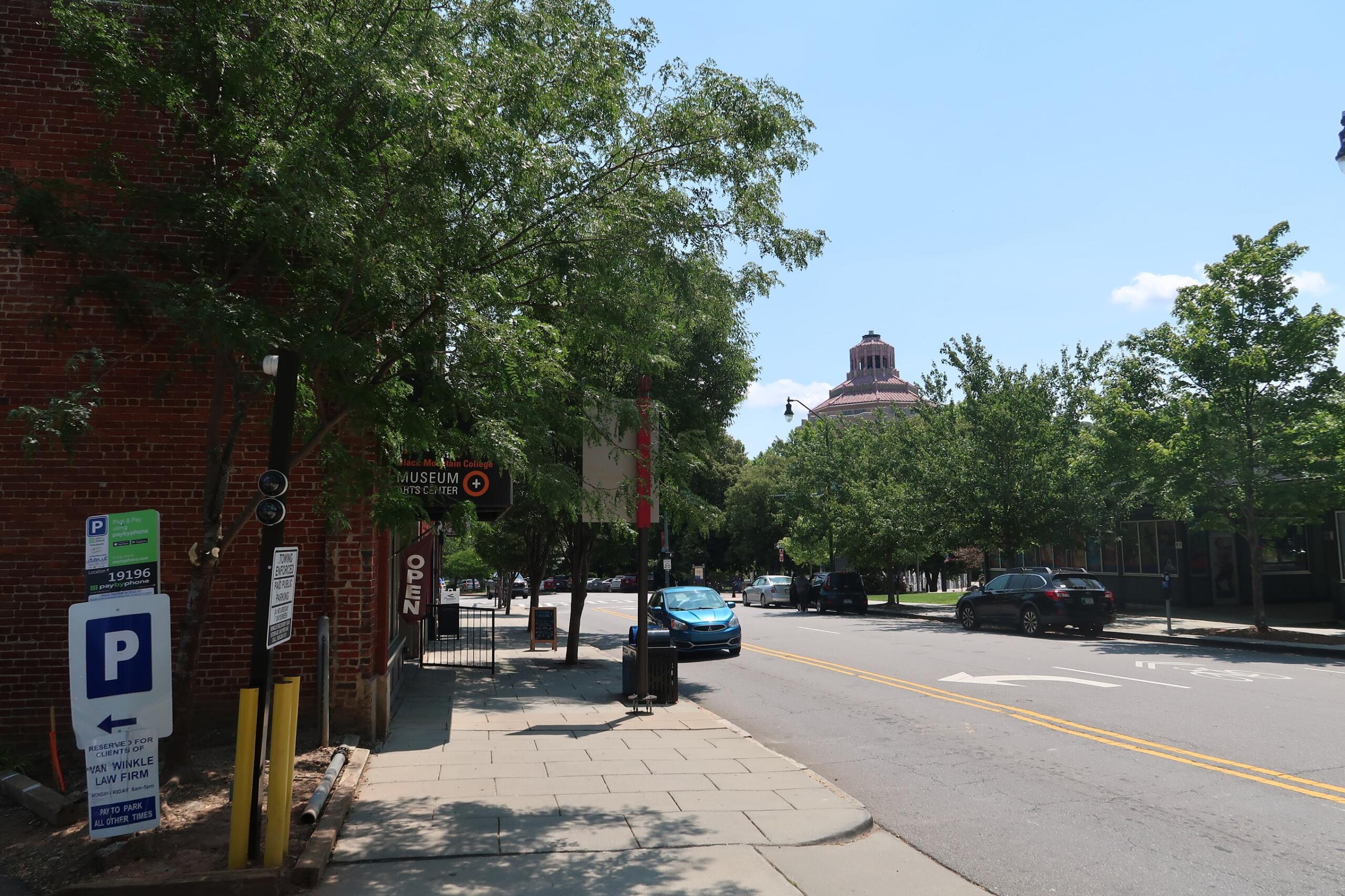 Street scene with a brick building and leafy trees, a sidewalk, and a distant domed building under a blue sky; cars parked along the street.