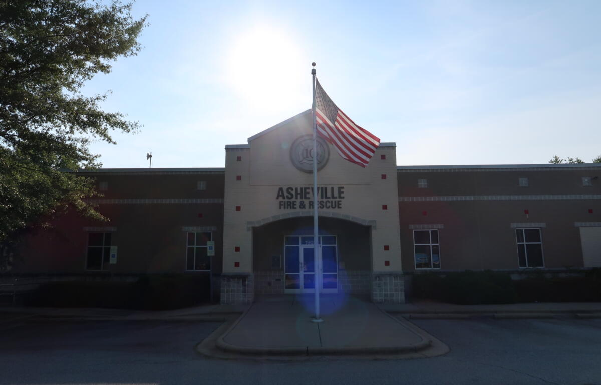 Front of Asheville Fire & Rescue station with an American flag on a flagpole and a bright sun flare over the roof