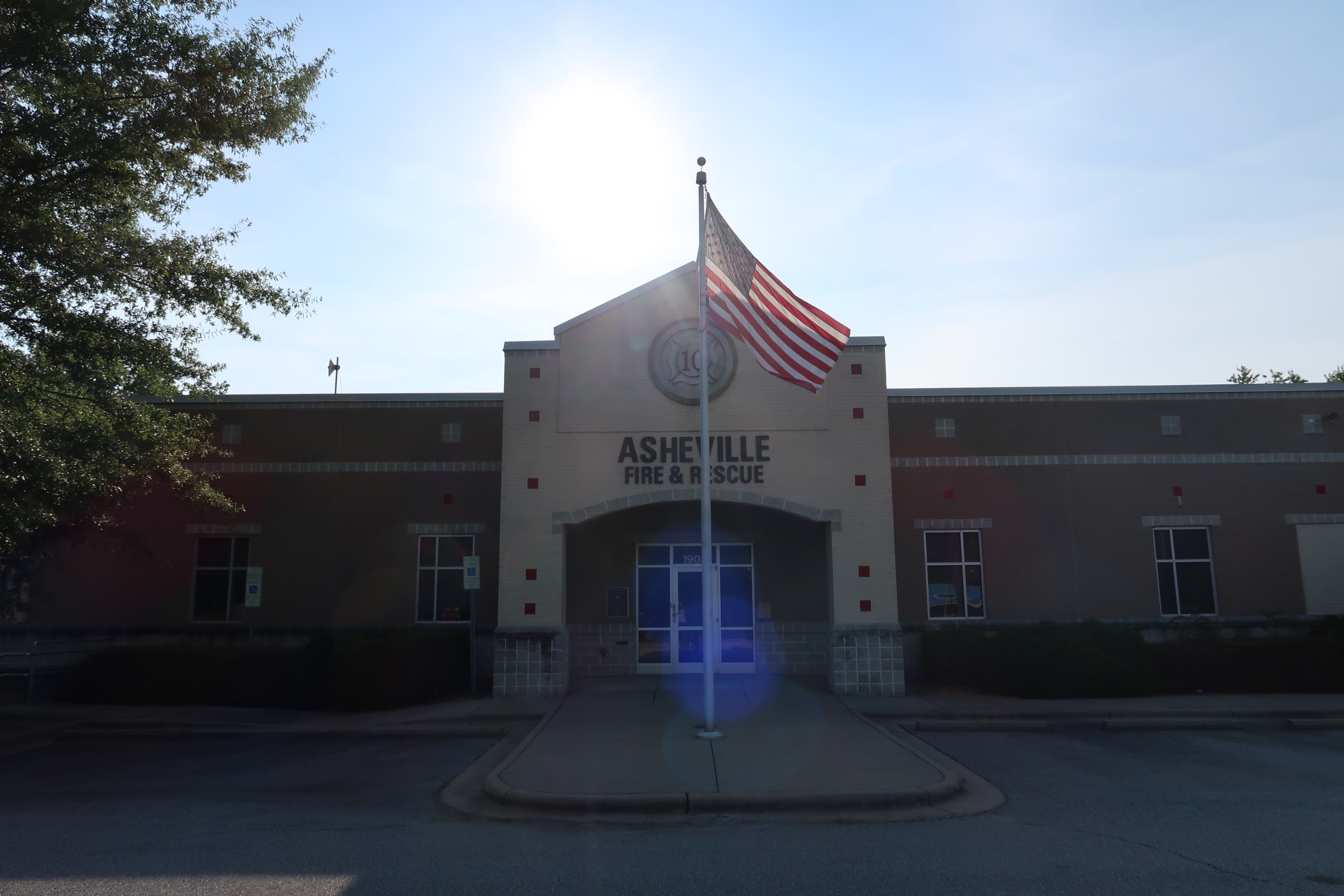 Front of Asheville Fire & Rescue station with an American flag on a flagpole and a bright sun flare over the roof
