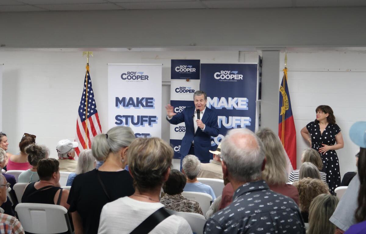 Speaker in a blue suit addresses an indoor political rally, with Roy Cooper banners and American flags behind him.